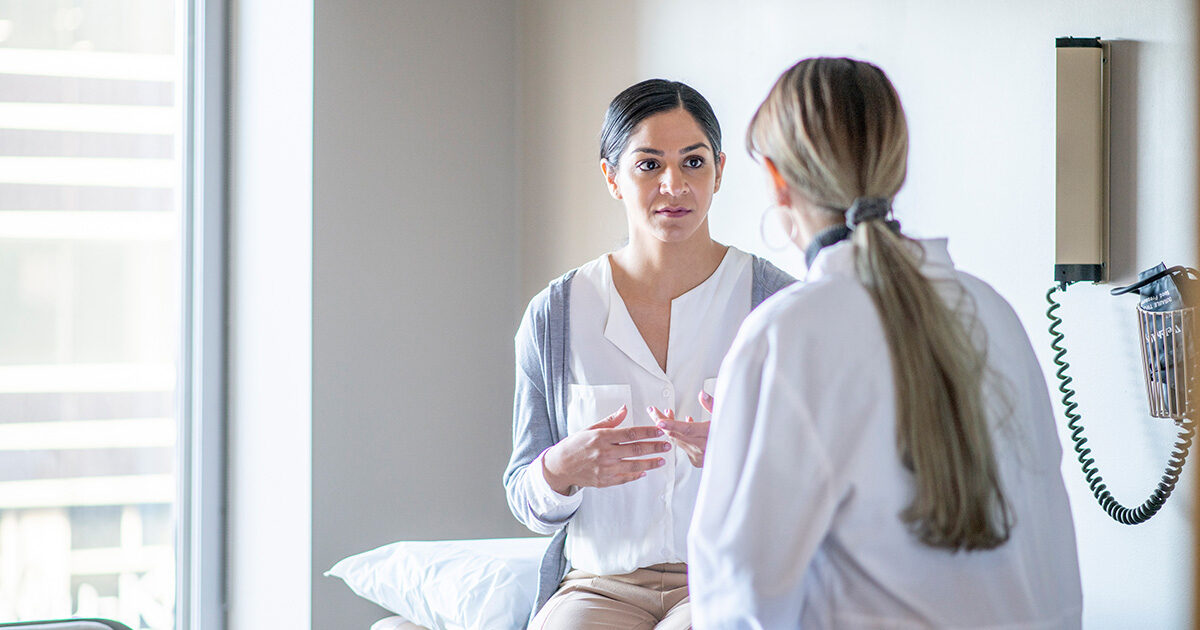 A woman sits upright on a medical exam table, speaking to another woman in a white coat.