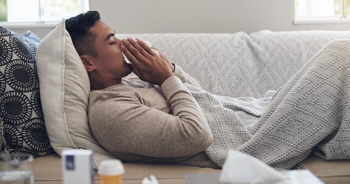 A young man lying on a couch, holding a tissue to his nose and mouth. He is covered in a throw blanket and medicine and tissues are visible on a coffee table in the foreground.