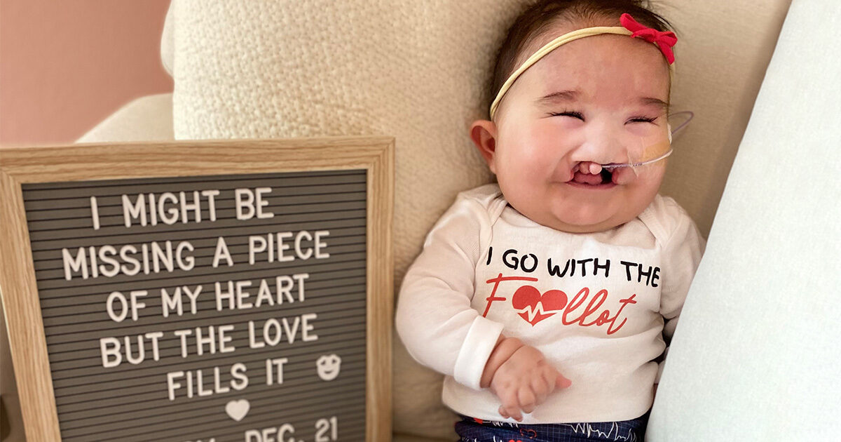 Baby Ruby Sedillo posed next to a gray letter board that reads "I might be missing a piece of my heart but the love fills it."  Her shirt says "I go with the Fallot", her pants are printed with hearts, and a red bow is attached to her headband