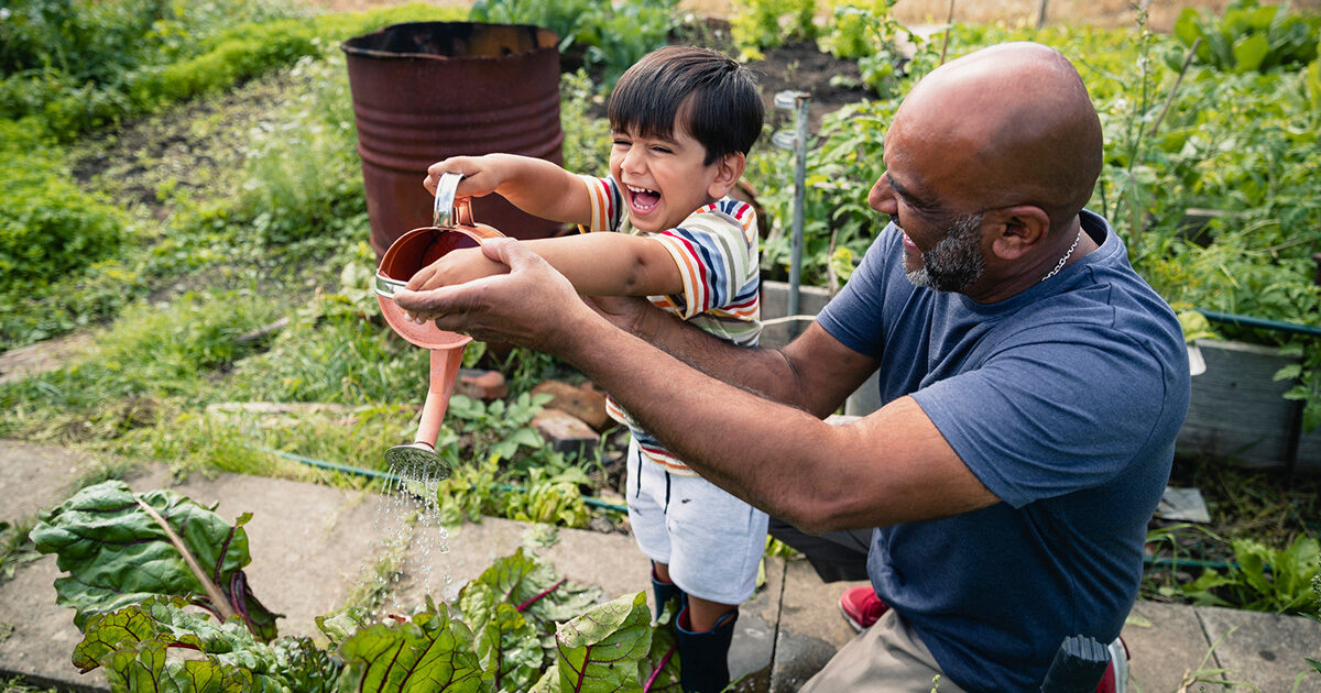 A man kneeling to help his son use a watering can on leafy plants.