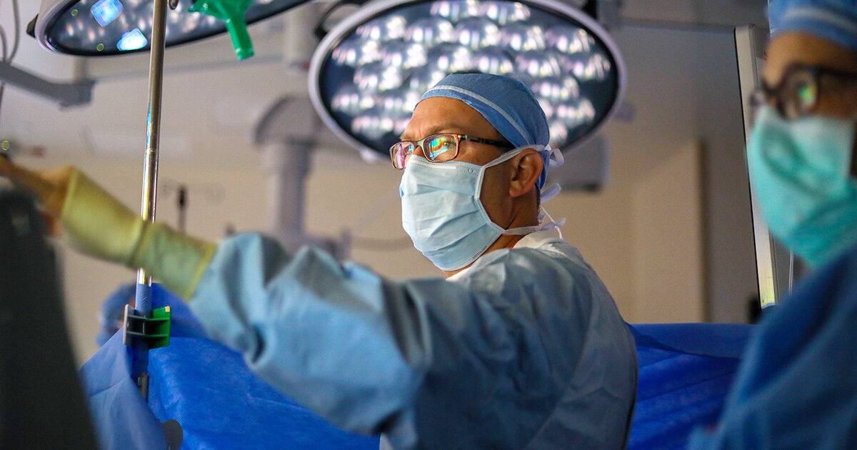 A surgeon wearing blue scrubs and a surgeon's cap is looking off at a screen in the OR.