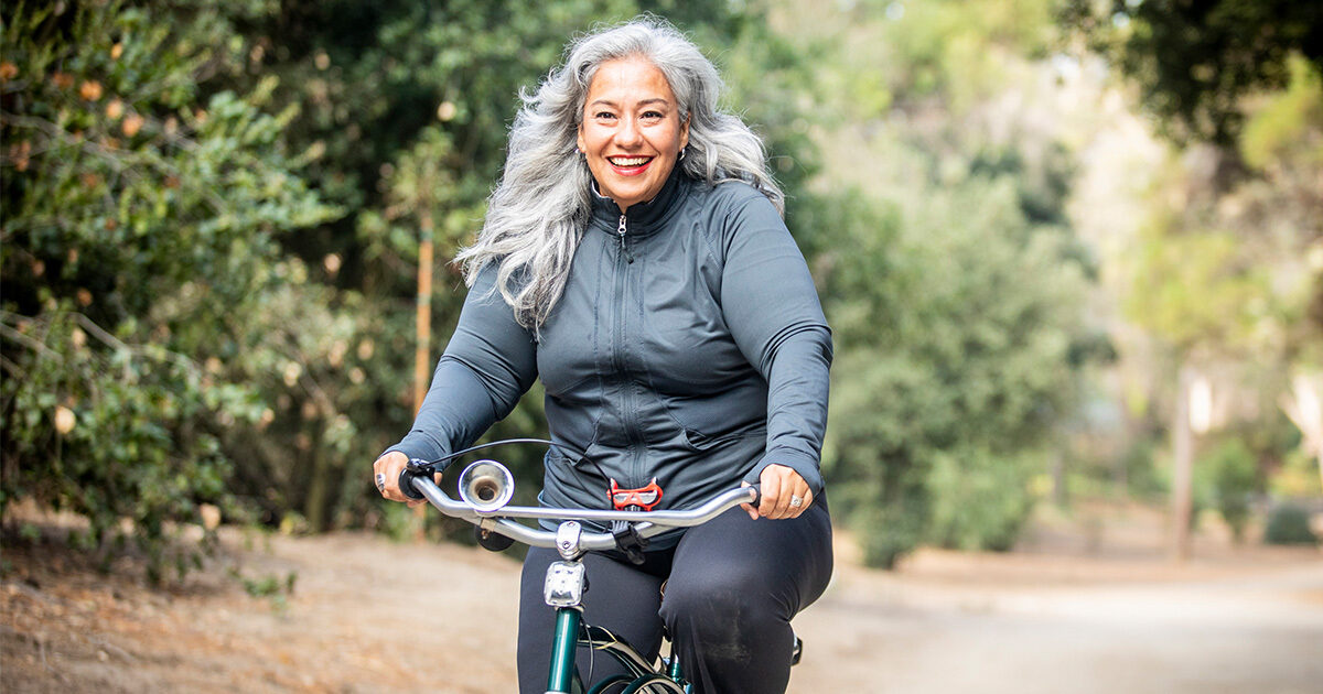 A smiling woman with gray hair rides a bike on a path lined with trees.