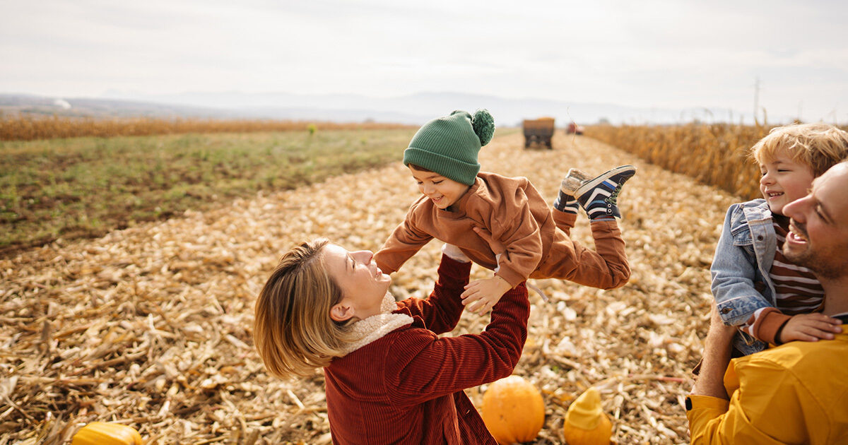 A woman lifts her child in the air in a pumpkin patch while her smiling husband holds their other child.