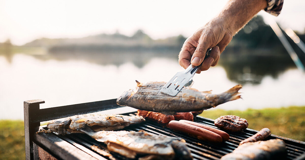 A closeup of whole fish on the grill. Someone is flipping one of the fish over with tongs, and a blurry lake is seen in the distance.