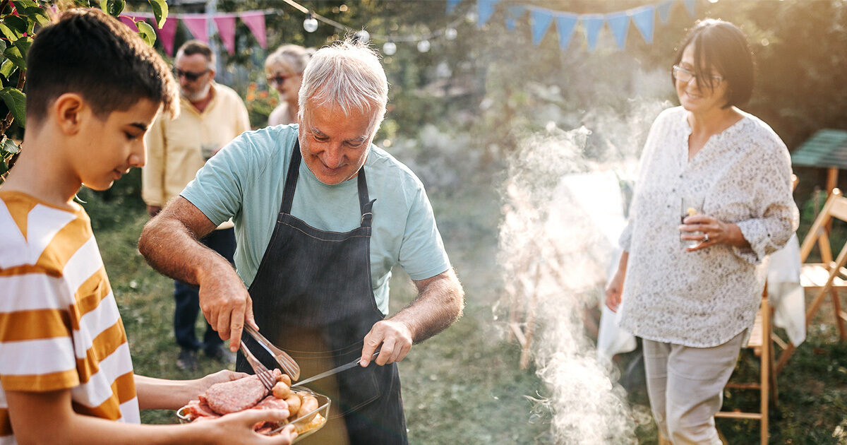 An older man wearing a black apron and a green shirt hands a younger kid a plate of barbecued food.  Another middle aged woman looks on and smiles. They're standing around a grill at an outdoor backyard party.