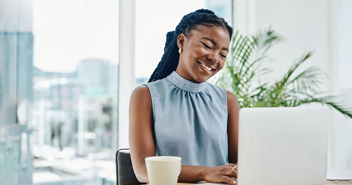 A young woman in a blue shirt tying at a laptop at her desk with a cup of coffee nearby. Her environment is brightly lit by a large window.