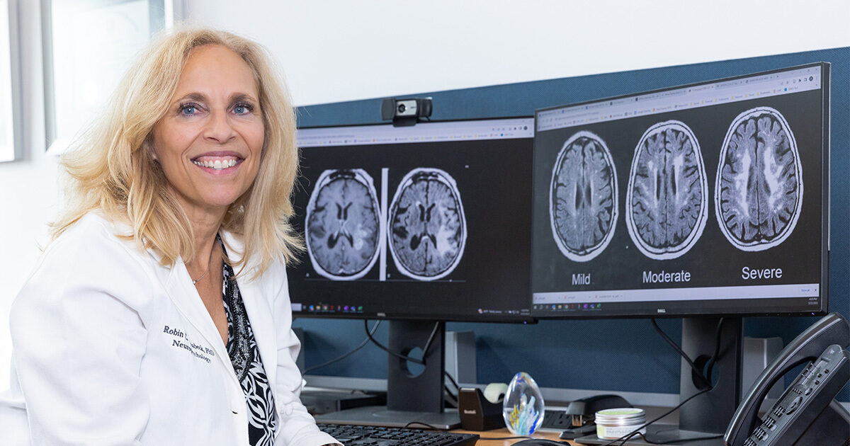 Robin Hilsabeck, PhD, ABPP, seated in front of two computer monitors displaying MRI brain scans.