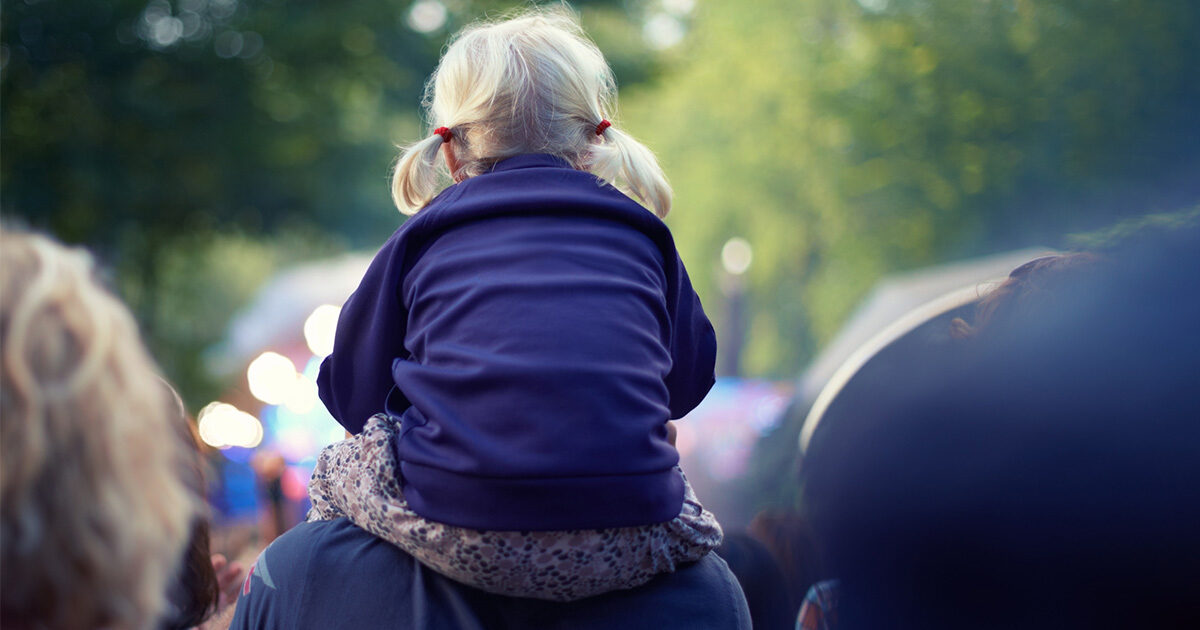 A little girl with light blonde pigtails sits on her dad's shoulders at an outdoor festival.