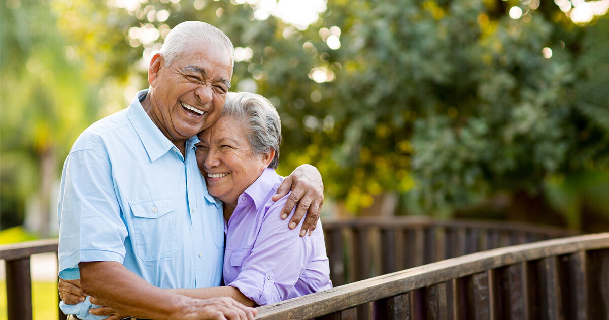 An older couple embraces on a wooden porch outdoors with trees in the background.