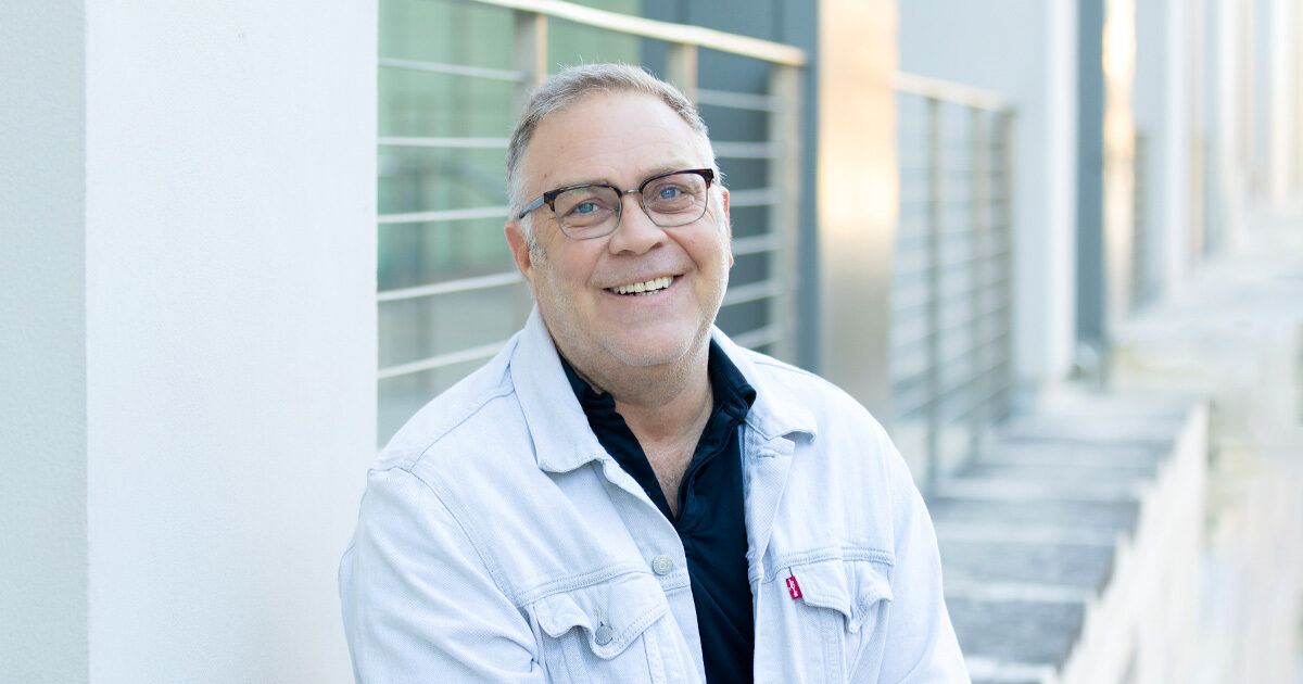 Jason Dudey smiling outside the Health Transformation Building at UT Health Austin. He is wearing a denim jacket and dark polo.