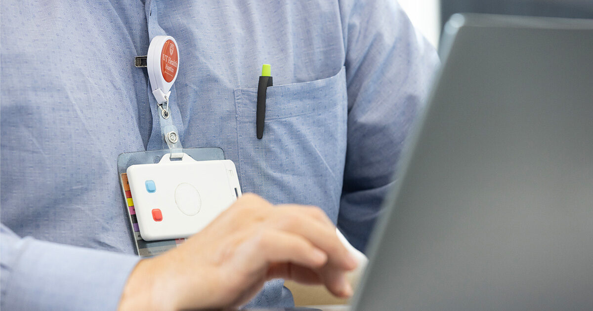 White wearable device hanging off a physician's badge holder, which is emblazoned with the UT Health Austin logo.