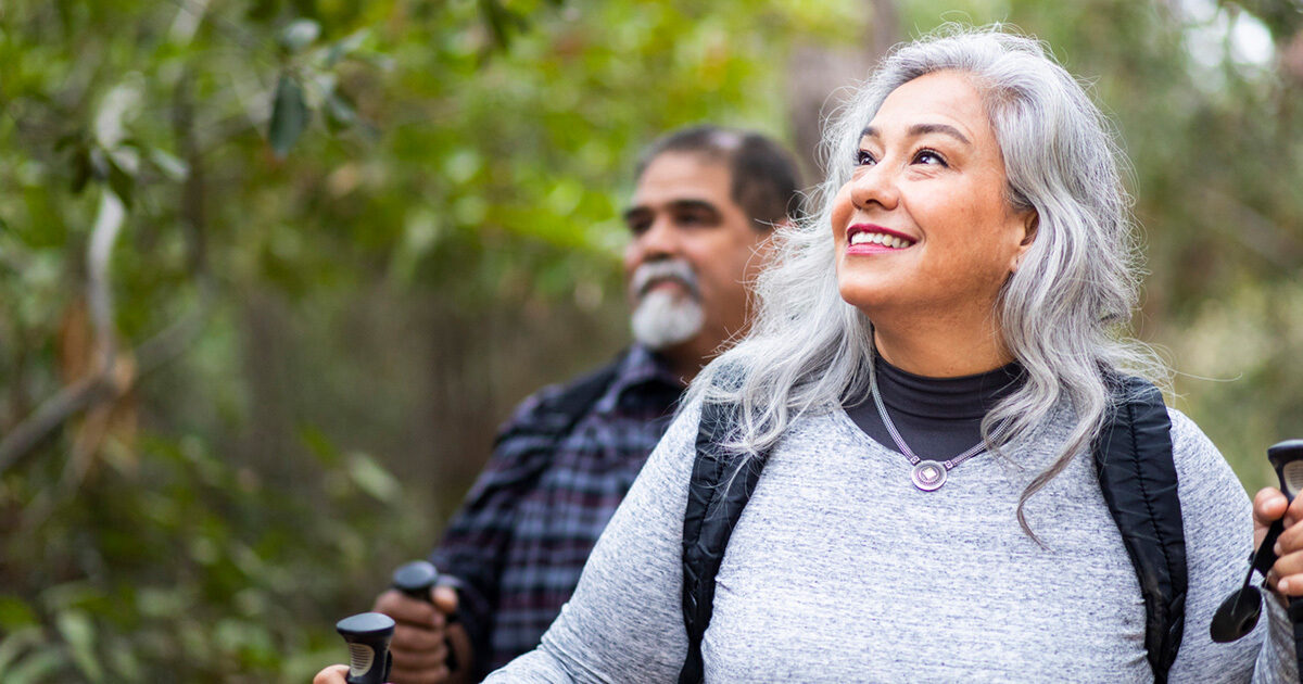 A woman with gray hair using walking sticks to hike through a forest. Behind her is a man with dark hair and a gray beard.