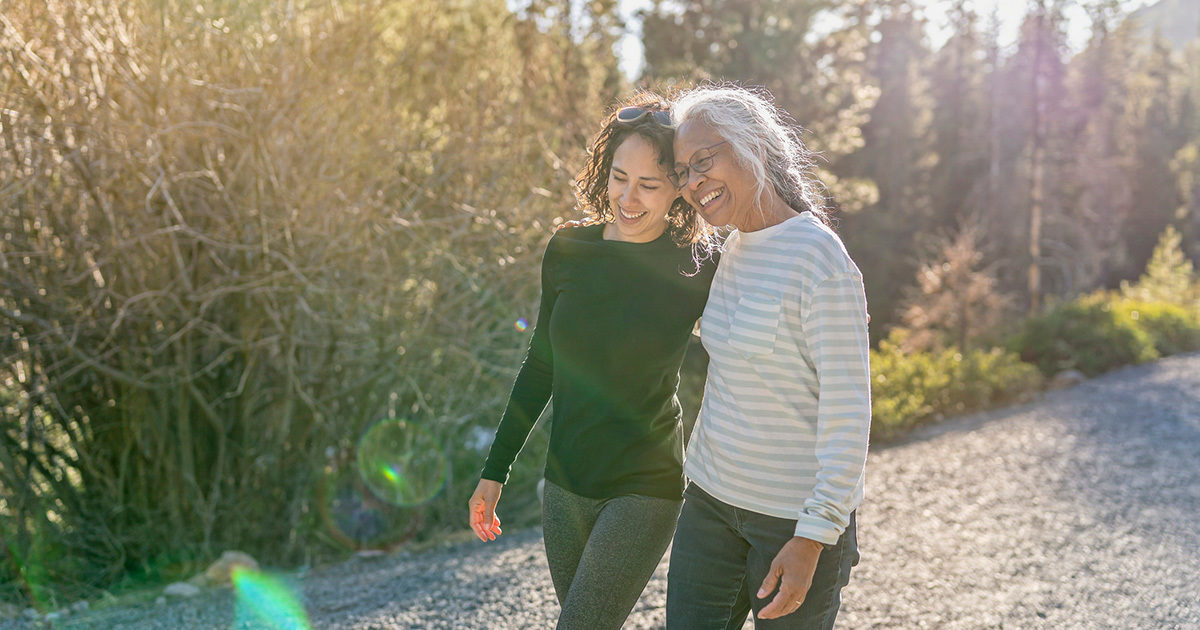 A woman and her retirement age mother walk arm in arm on a hiking trail surrounded by trees on a sunny day.