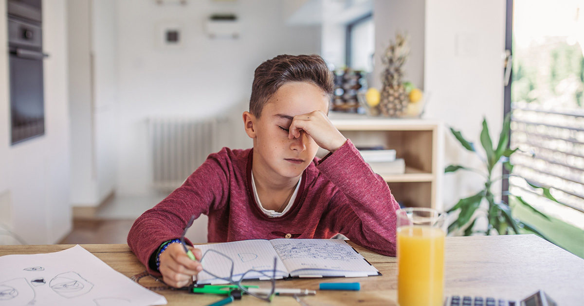 A young male student, with glasses in his right hand, studying a book, rests his head on his left hand.