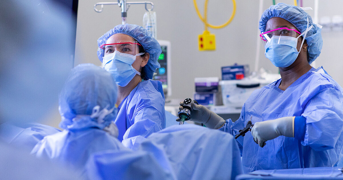 A group of clinicians wear surgical gowns, masks, hairnets, and protective eyewear in the operating room.