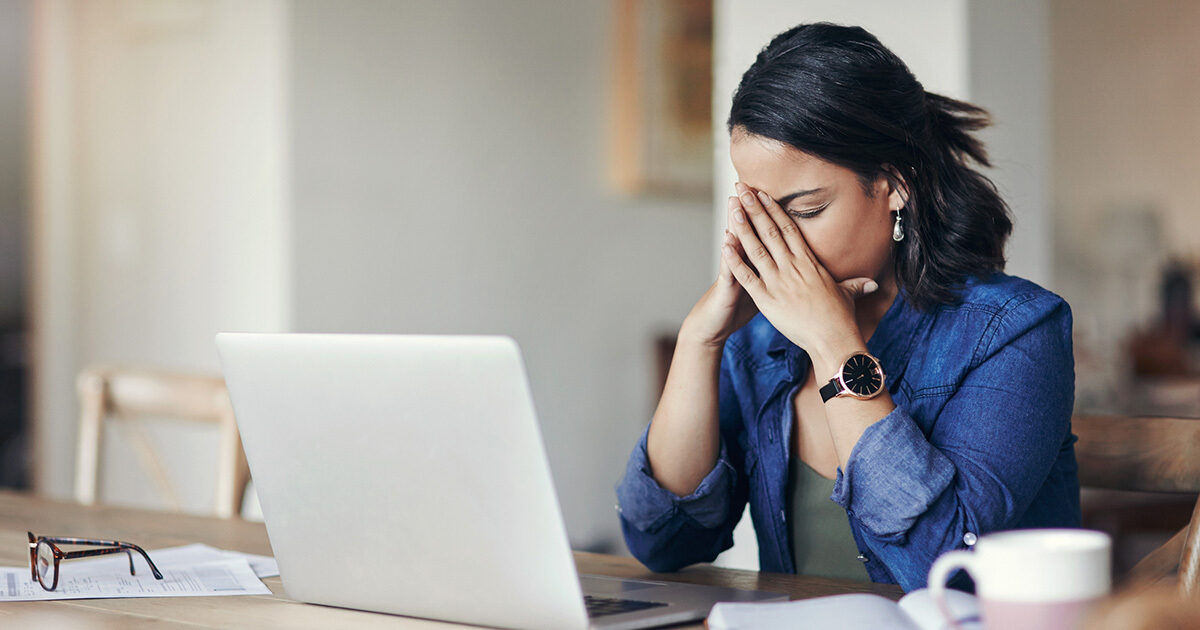 A woman sitting at a table with a laptop cradles her face in her hands.