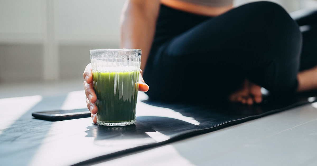 A close up of a glass of a green smoothie, being held by a woman seated on a yoga mat.