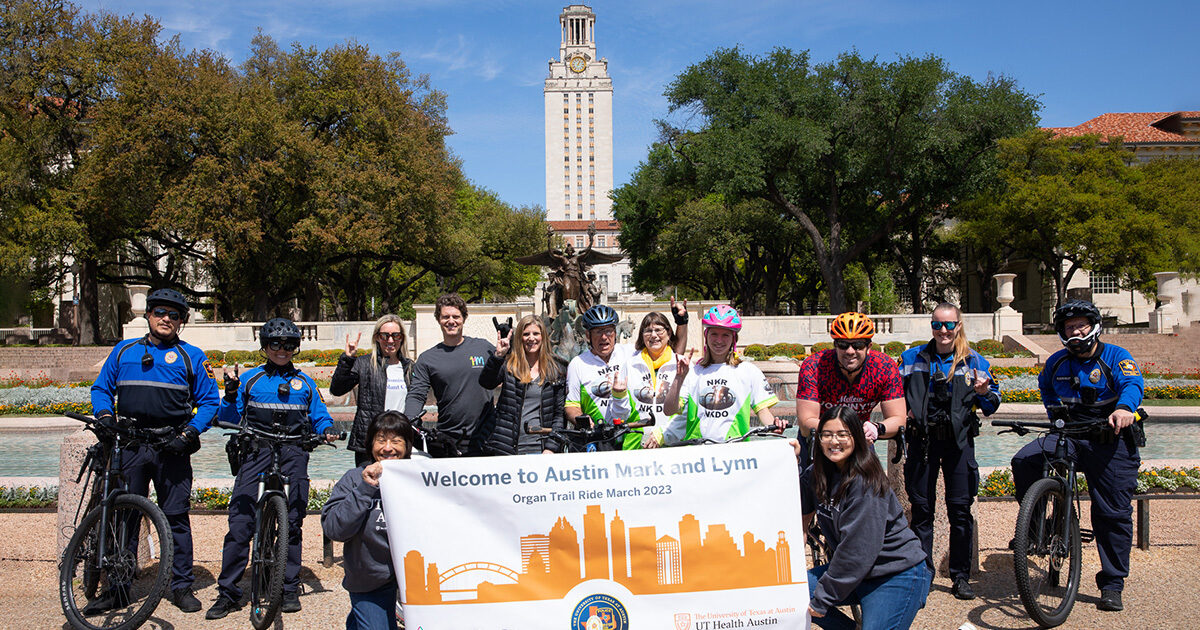 Participants in the Organ Trail ride through the UT Austin campus posing in front of Littlefield Fountain with a banner reading "Welcome to Austin, Mark and Lynn. Organ Trail March 2023." The UT tower is visible in the background.