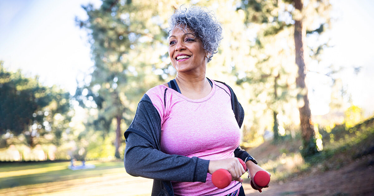 A short-haired woman in exercise clothes carrying weights as she walks through a forest.