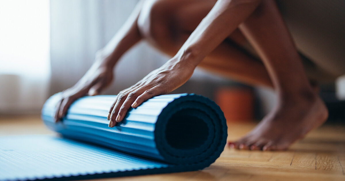 A closeup of a dark complected woman's hands are seen rolling out a blue yoga mat.  She is indoors on a hardwood floor with soft natural light filtering in through the window.