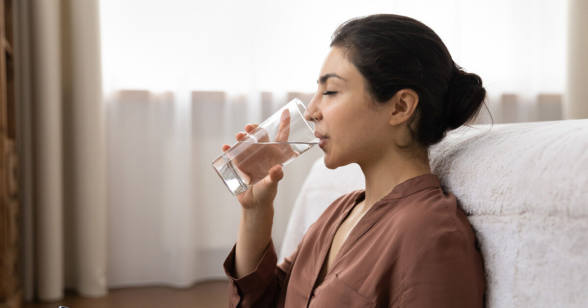 A woman sits on the floor of her bedroom, leans against her bed, and drinks from a glass of water.