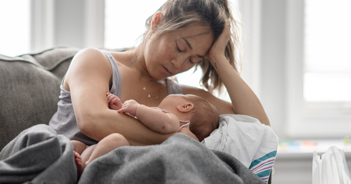 A woman on seated on a couch cradling a baby in one arm and resting her own head in the other hand.