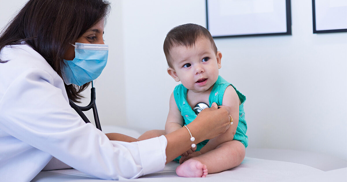 A female doctor in a white coat wearing a surgical mask listens to an infant's heartbeat using a stethoscope.