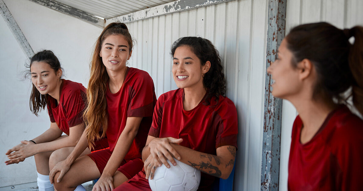 Three teenage girls in red soccer jerseys sit in the shade of a dugout and talk amongst each other.