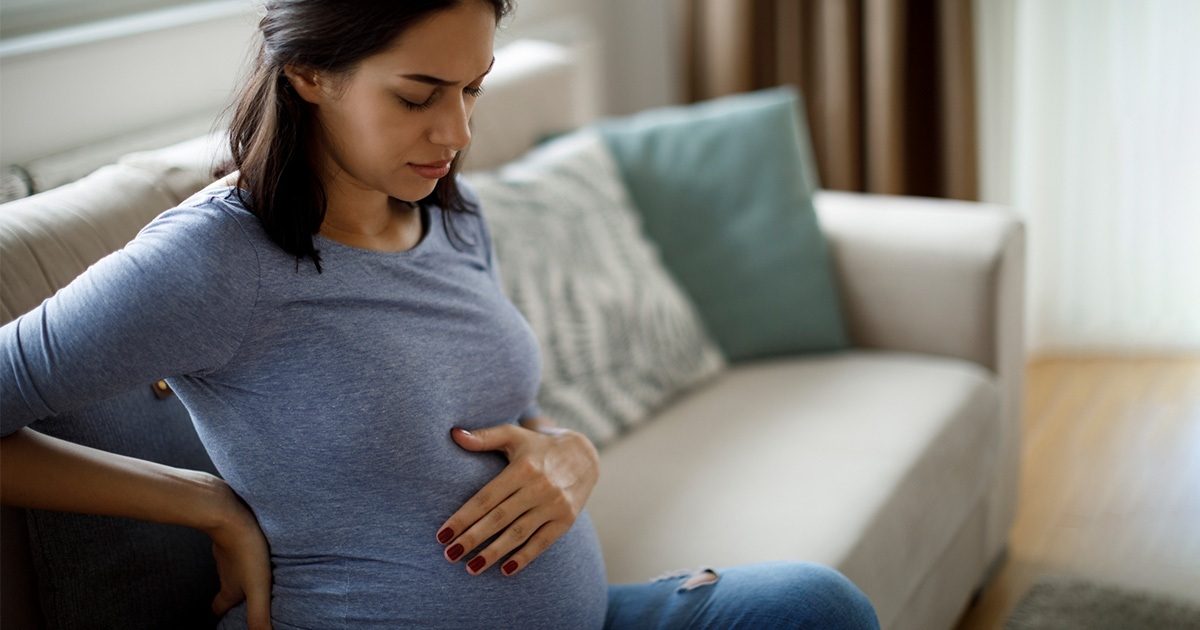 A pregnant woman is sitting on a white couch. She's wearing a blue shirt, looking down, and has one hand on her back and the other on her baby bump.