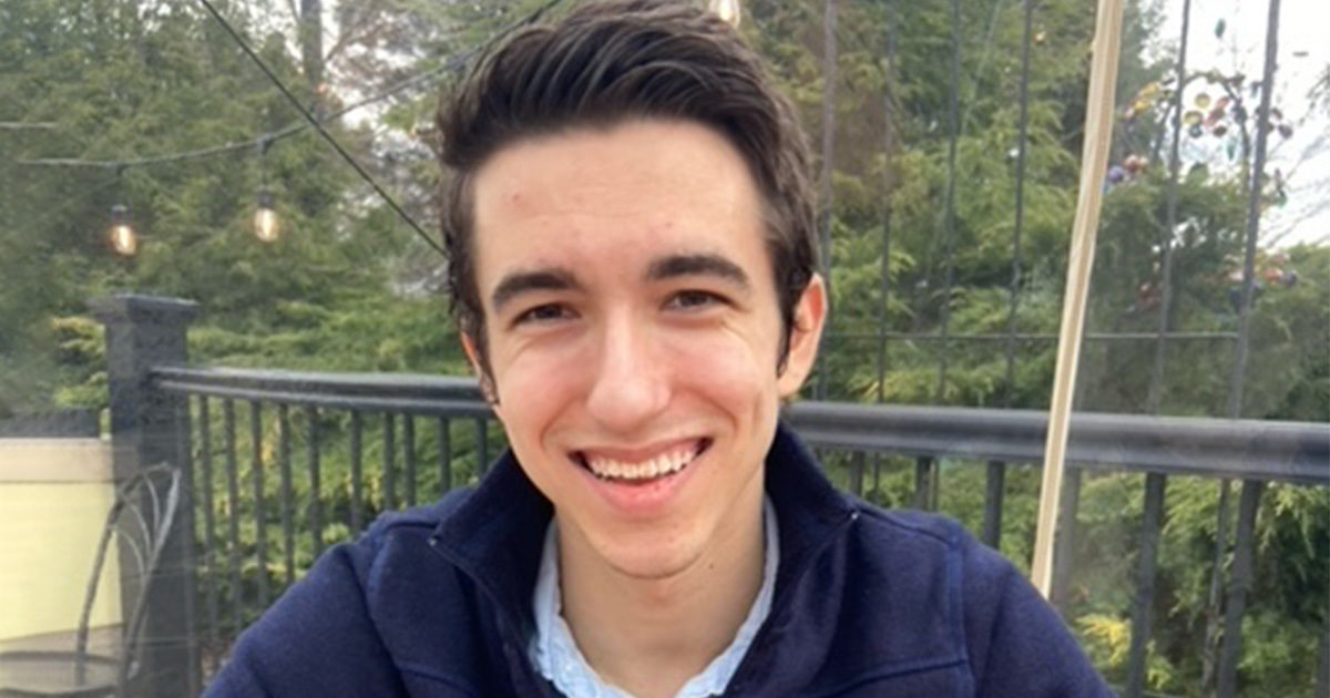 Dom Rainelli is wearing a blue jacket, looking at the camera, and smiling. He is sitting outside on a deck on an overcast day.