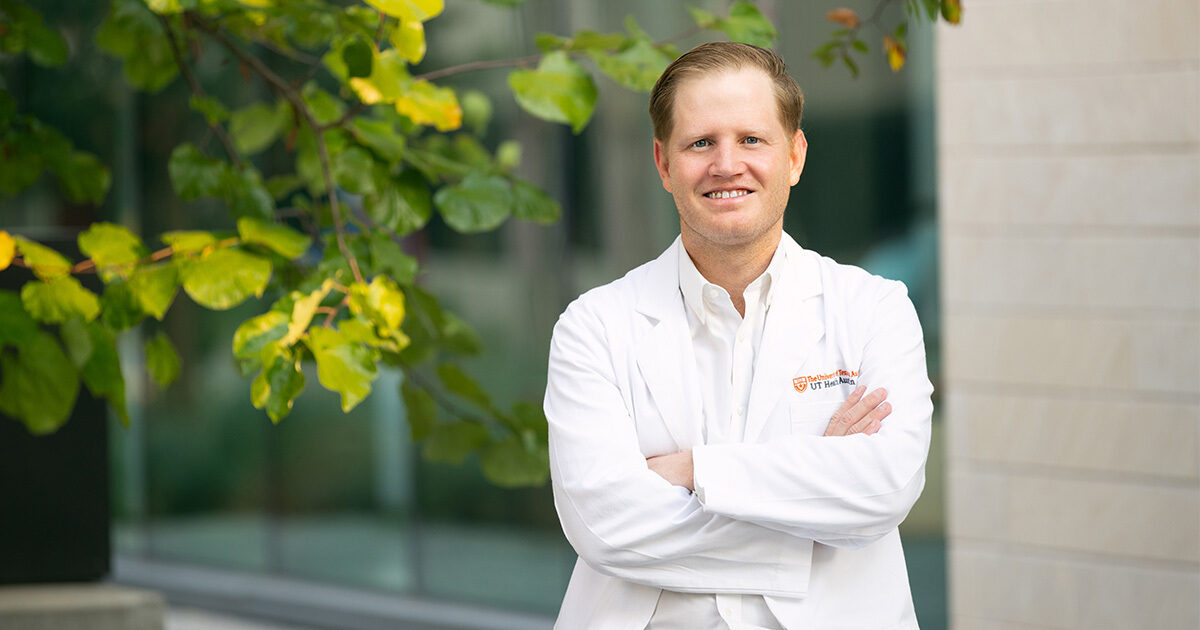 Endocrine surgeon Samuel E. "Rhett" Long, MD, FACS, posing next to a tree outside of the Health Discovery Building in downtown Austin. His arms are crossed and he is wearing a white coat with an embroidered UT Health Austin logo.