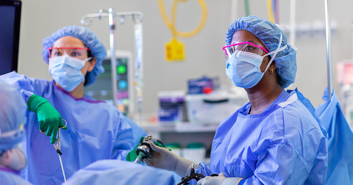 Two clinicians wear surgical gowns, masks, hairnets, and protective eyewear in the operating room.