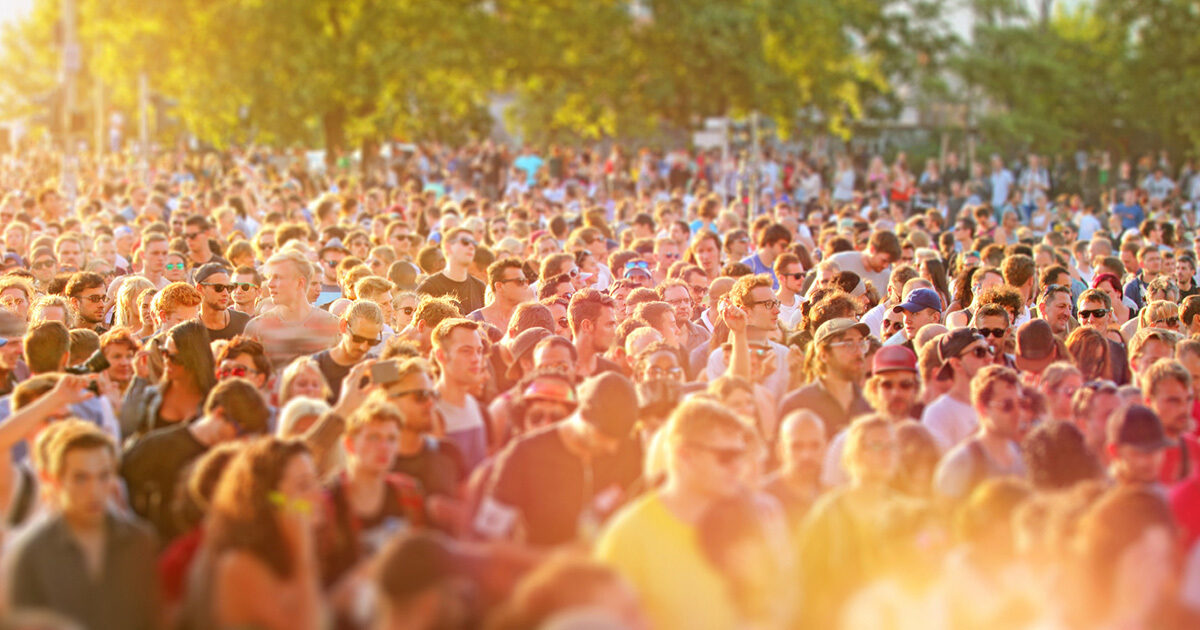 A large crowd gathers outside at a music festival. The warm sunlight is filtering through the trees.