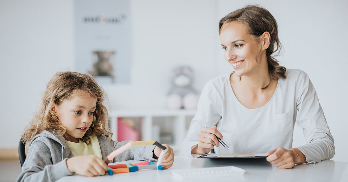 A young female child, working on a puzzle, and an adult woman, holding a tablet, are seated at a desk in a classroom setting.