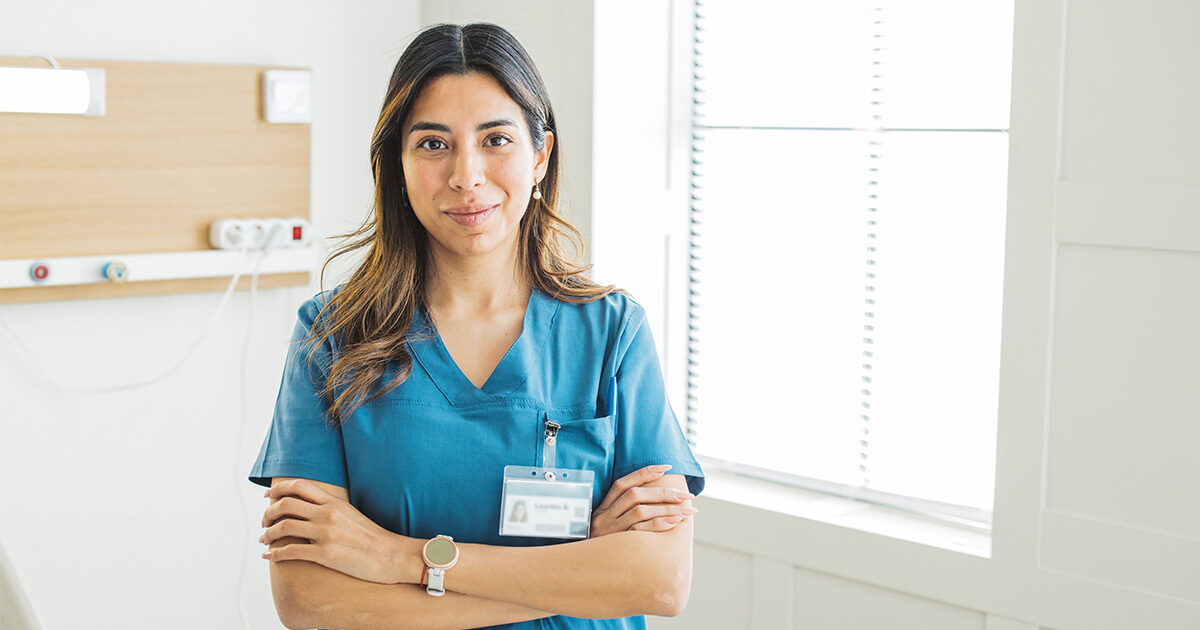 A long-haired woman with teal scrubs and a name badge crossing her arms and smiling near a window.