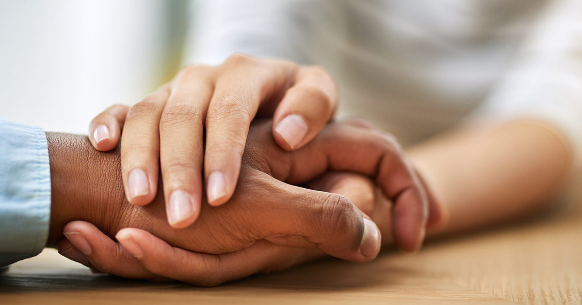 A close up of a young woman's hands enclosed around a Black man's hand in a comforting gesture.