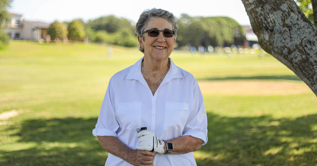 Chris, a knee patient at US Health Austin, stands smiling on a golf course, her hands wrapped around the handle of a golf club.