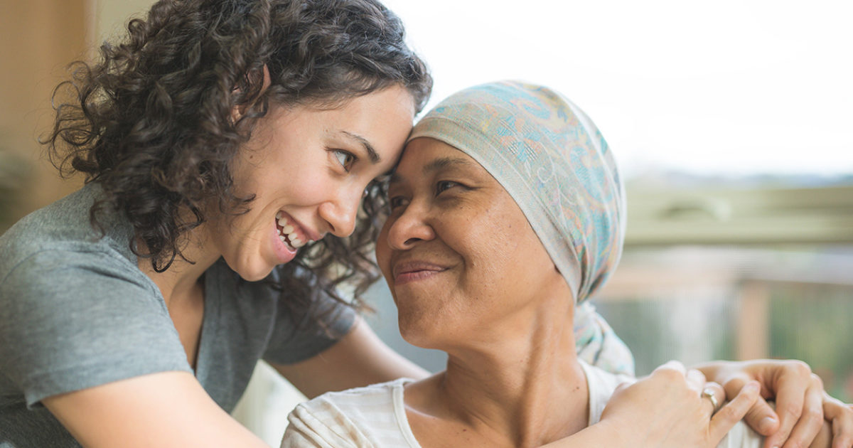 A young woman smiles at and embraces an older woman wearing a headscarf who is seated and looking at the younger woman.