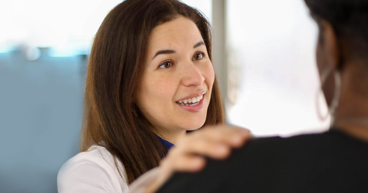Christina Salazar, MD, FACOG, Minimally Invasive Gynecologic Surgeon with the Women’s Health clinic, greets a patient. Dr. Salazar's hand is on the woman's shoulder and she is smiling.