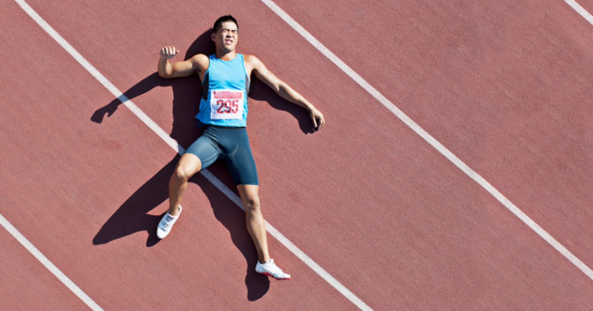 A top-down view of a male track and field athlete laying on his back on a running track. His face is contorted in a grimace of discomfort.