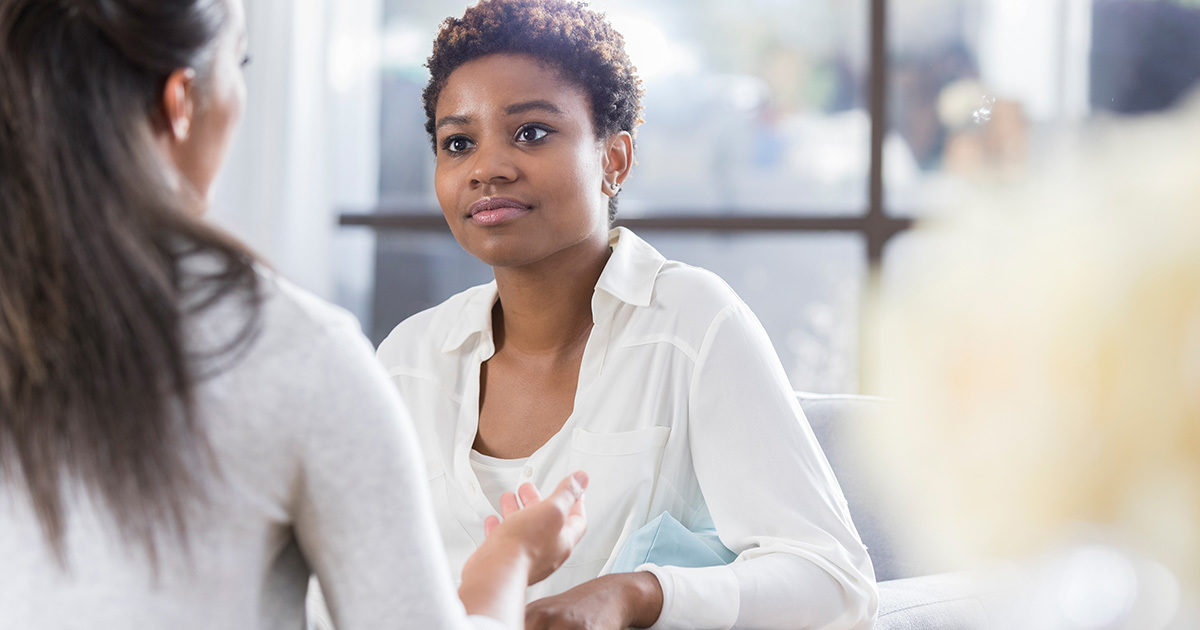 A young African-American woman in a white shirt listens as another woman speaks to her.