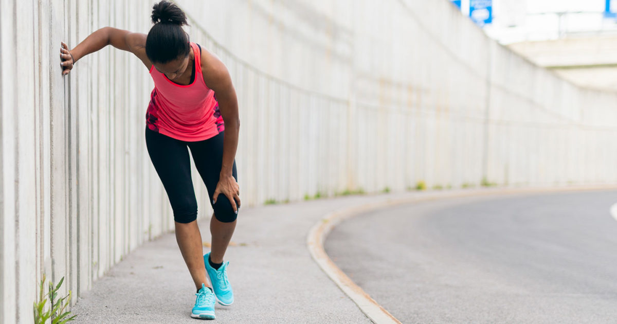 A woman in athletic gear, stops on the side of the road holding on to the wall with her right hand, while she leans over to hold her knee with her left hand.