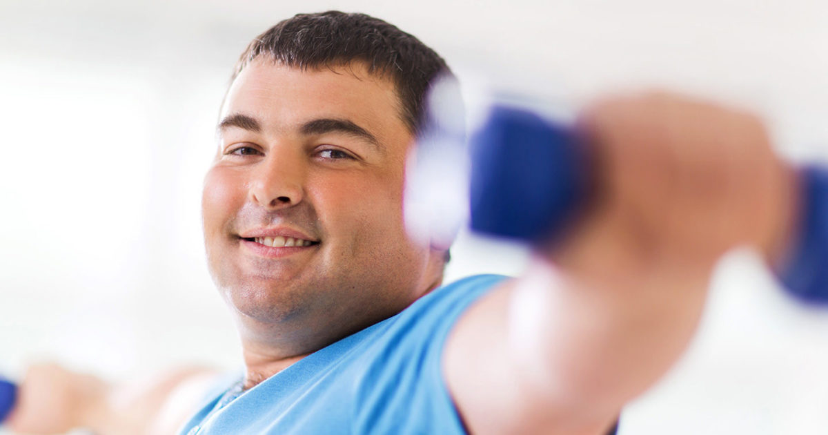 A man smiles at the camera as he lifts a pair of dumbbells.