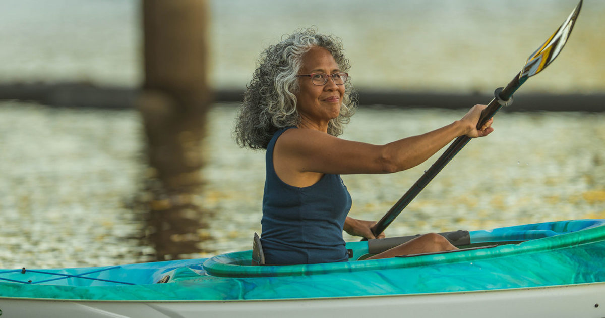 A mature woman with gray curly hair wearing glasses, paddles a kayak near a pier.