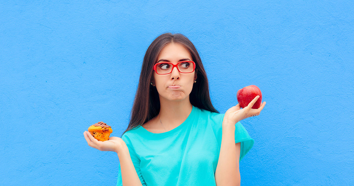 A young woman stands in front of a bright blue background holding an apple in one hand and a muffin in the other. She wears an expression of thoughtful confusion.