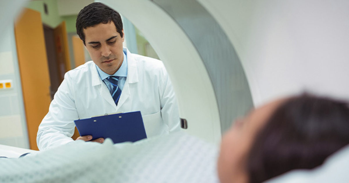 In this photo, taken from inside an MRI scan machine, a male clinician peers at a clipboard while a female patient waits inside the scanner.