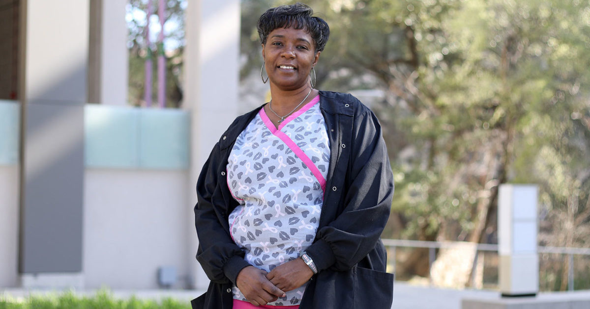 Patrice, a patient in Women's Health, stands outdoors with her hands clasped in front of her hips, smiling.