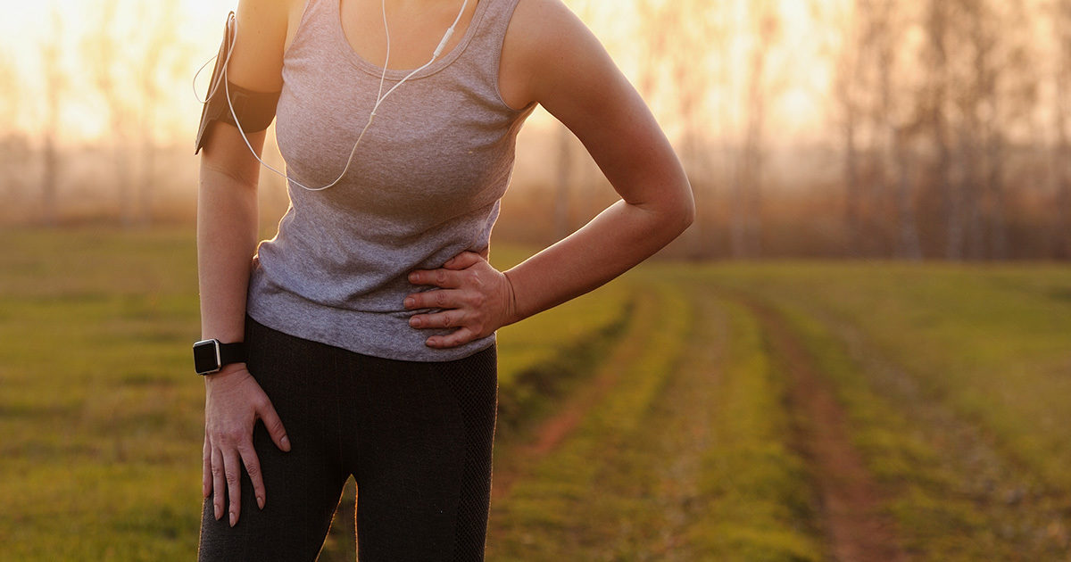 A closeup view of a woman wearing athletic clothes, standing outside, while she holds the side of her abdomen in an expression of discomfort.