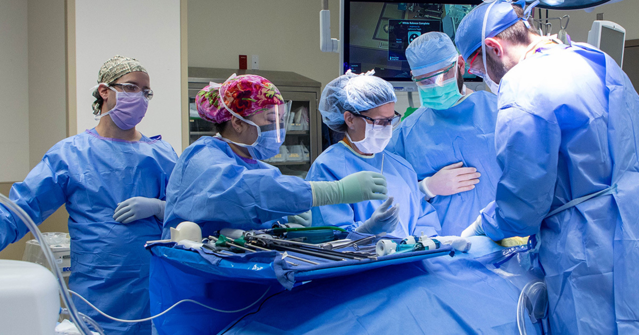 Dr. Nicole Turgeon and four members of her surgical team standing around a tray that holds several surgical instruments. All five providers are wearing surgical caps, masks, gowns, and gloves.