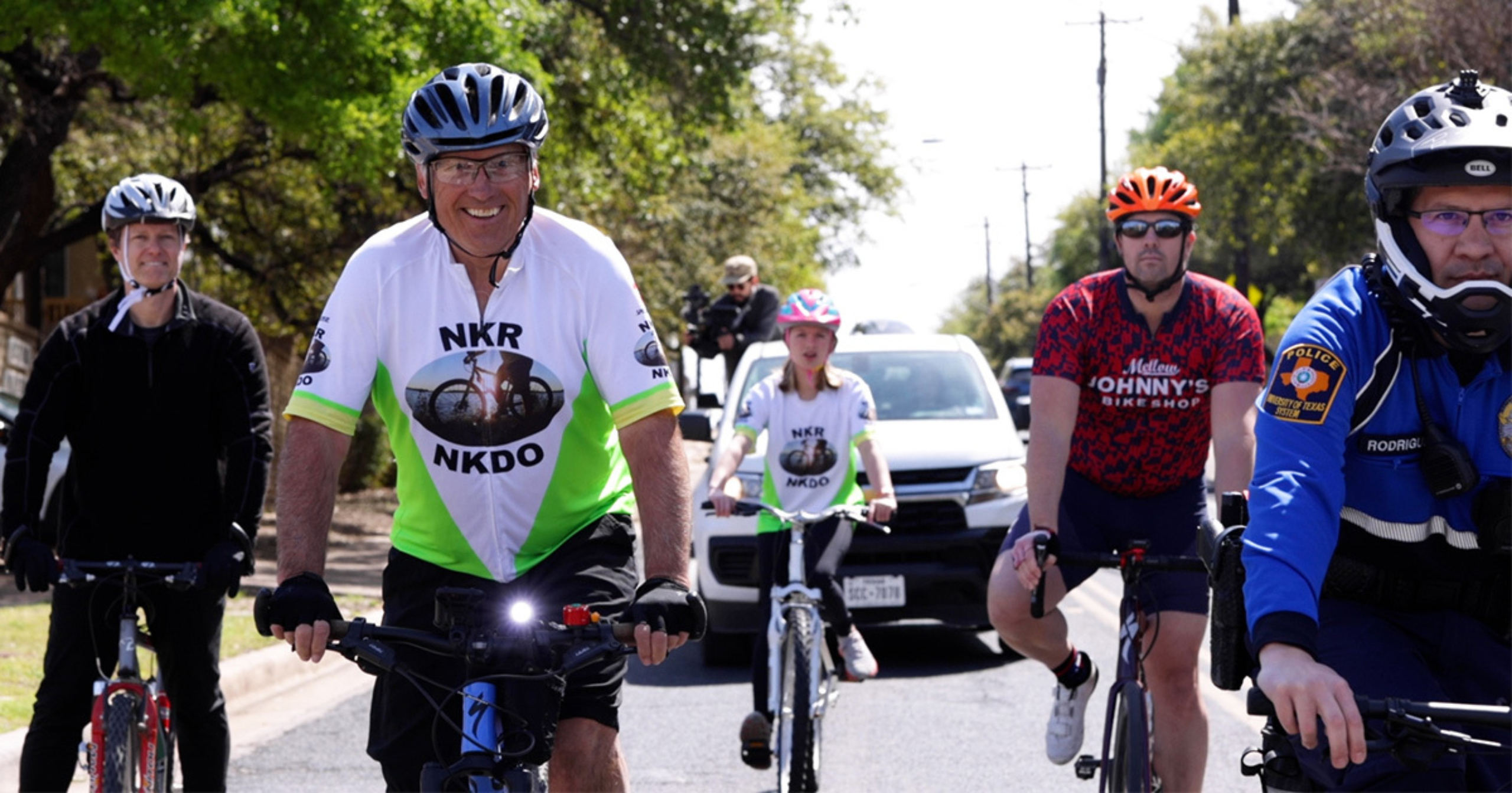 Travis Watson, Mark Scotch, Rowan Sebek, Chase Sebek and an officer from the University of Texas Police Department riding their bikes down a road lined with trees. Behind them is a man in a truck with a camera.